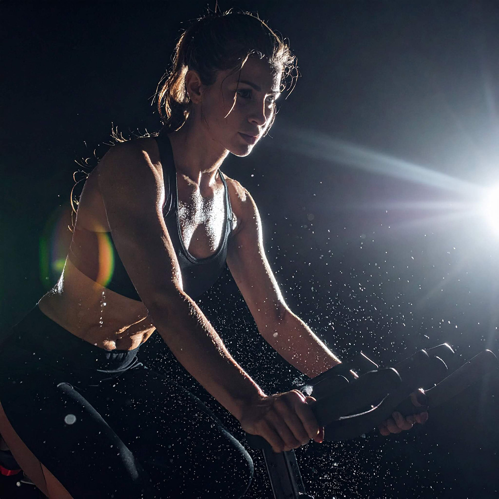 Firefly_暗闇のフィットネススタジオで、女性がエクササイズバイクを全力で漕いでいる。強いスポットライトが背後から当たり、飛び散る汗の粒が光に照らされて輝いている。背景(1)